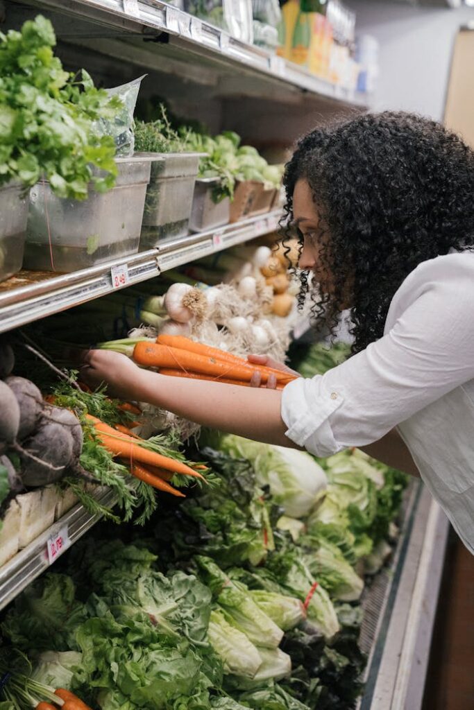 Woman selecting fresh organic carrots from a grocery store shelf filled with vegetables.