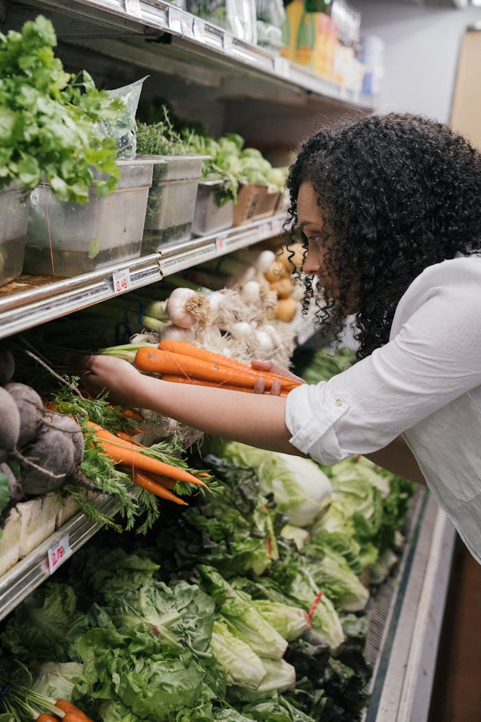Woman selecting fresh organic carrots from a grocery store shelf filled with vegetables.